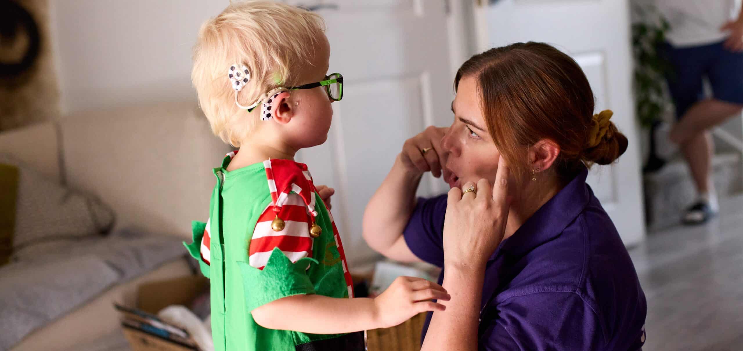 Harvey is stood in his elf suit looking at Laura. Laura is sat in front of him pointing to her face.
