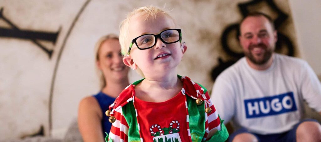 Harvey is stood smiling in his elf outfit. Kim and her partner are sat behind him.