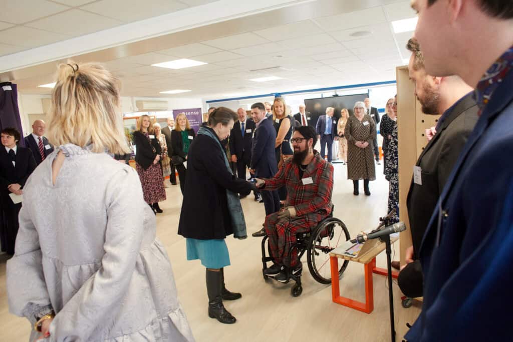 HRH The Princess Royal shakes hands with a man in a wheelchair wearing a grey and red checked suit in a room surrounded by people.