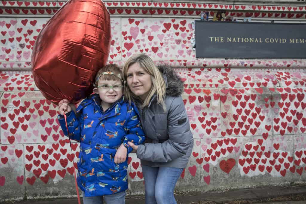 Landscape shot of the National Covid Memorial Wall covered in hand drawn red and pink hearts. The plaque on the wall reads 'The National Covid Memorial Wall'. A boy in a blue coat is facing the camera with his mum, a woman in a grey coat standing next to him. The boy hold a large heart shaped red balloon.