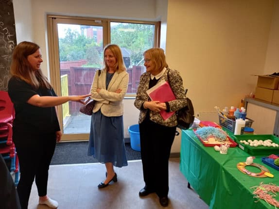 Three women standing talking. They stand next to a table with a green table cloth, covered in arts and crafts items used for sensory play