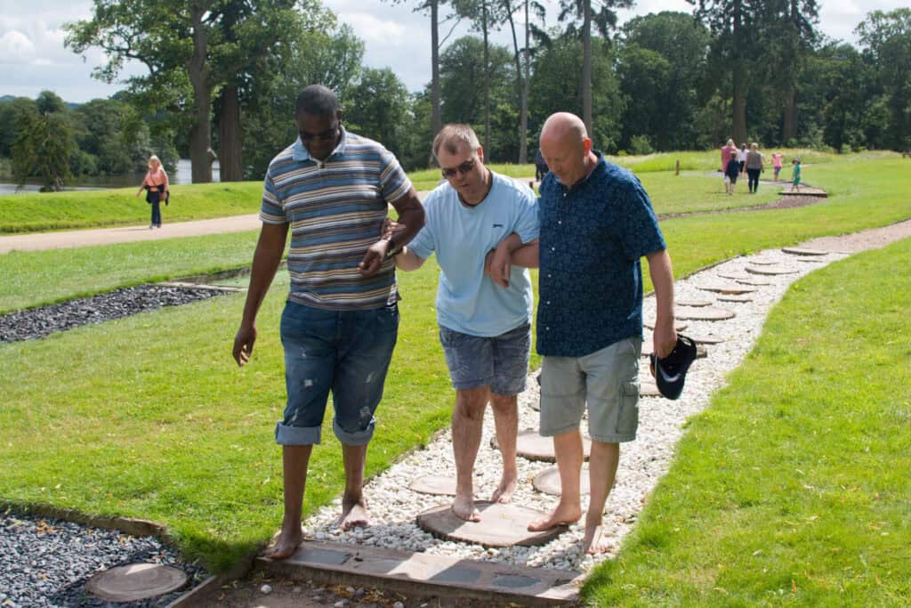 Two men guide another man along a path in a green garden.