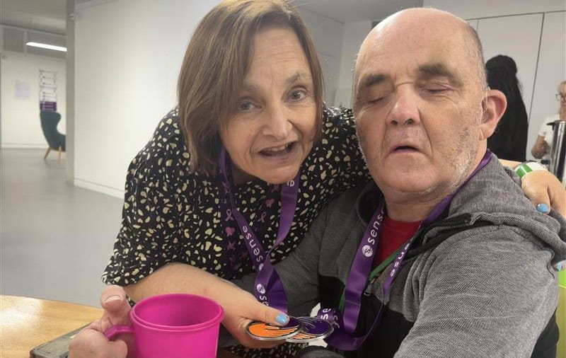 Joy is stood with her left around around Joe, who is sat down. They are both smiling at the camera. Joy is holding two Sense medals which in a ribbon around their necks.