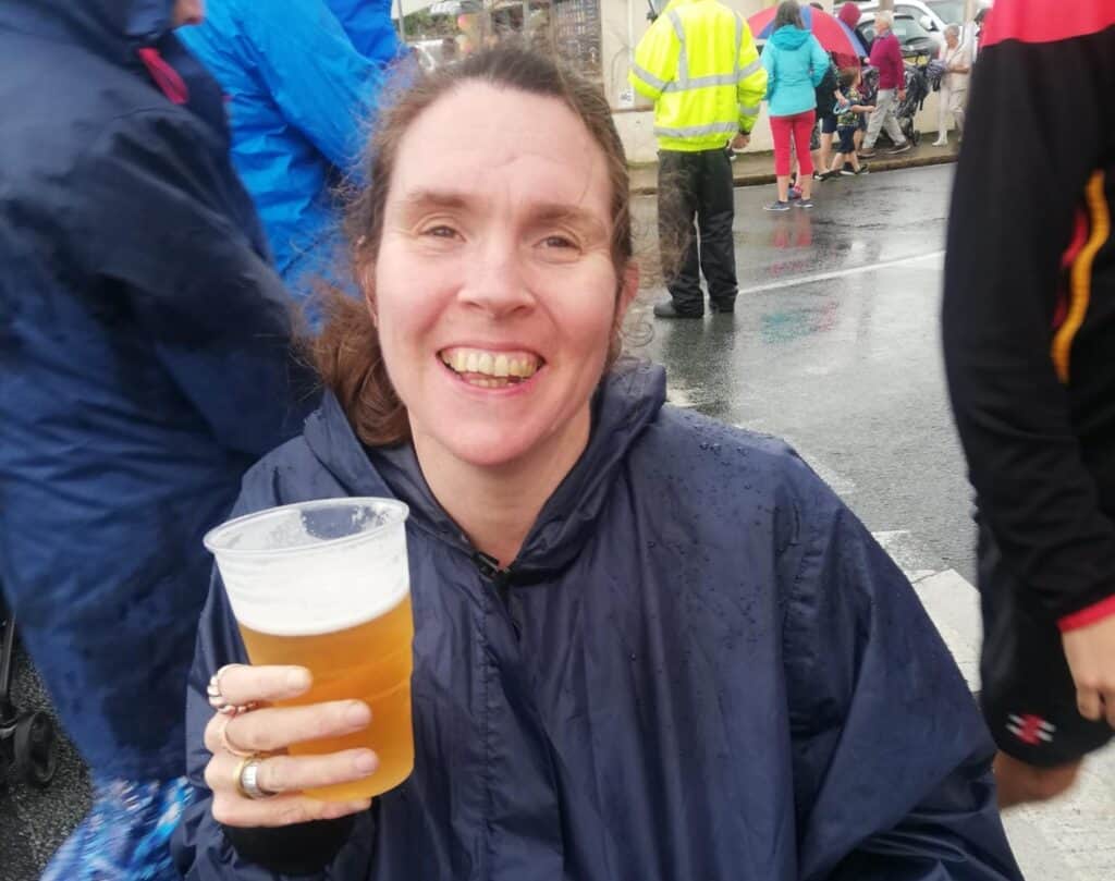 Joanna is sat in a dark blue poncho, smiling widely, holding a pint of lager.