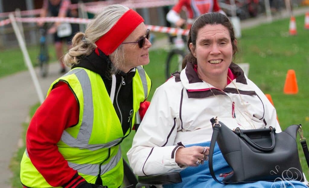 Joanna is sat in her wheelchair, with her black bag on her lap. There is a lady in a Hi-Vis vest helping to push her wheelchair from behind. They are in conversation.