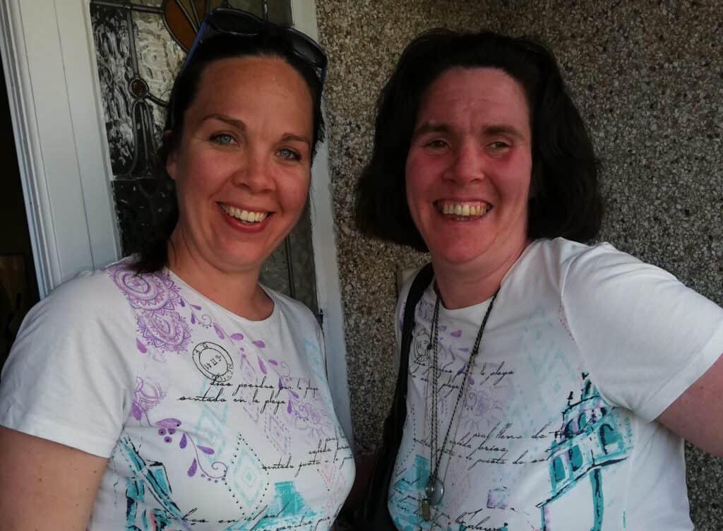 Berni and Joanna are stood side by side, smiling widely at the camera. They are wearing matching white t-shirts with colourful patterns and writing on.