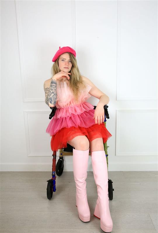 A woman with long blonde hair sitting on a mobility aid, wearing a pink dress with ots of ruffles, pale pink knee high boots and a bright pink beret