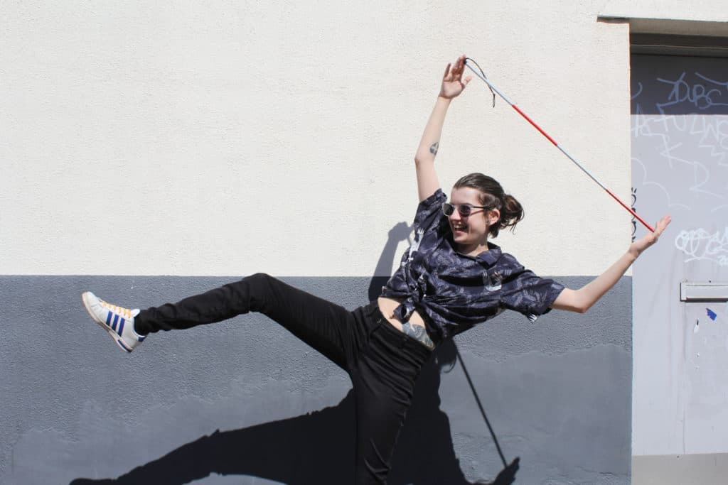 Outdoors, a person with a shirt and black jeans stands on one leg. Above their head, they're holding a white and red cane. They're smiling.