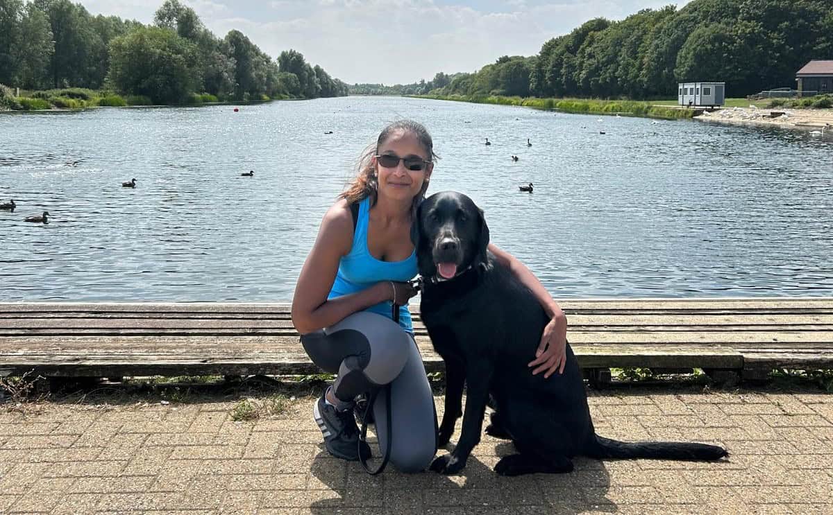 Jaina is kneeling next to her dog Dave, with her left arm around him. They are posing for a picture in front of a sunny river.