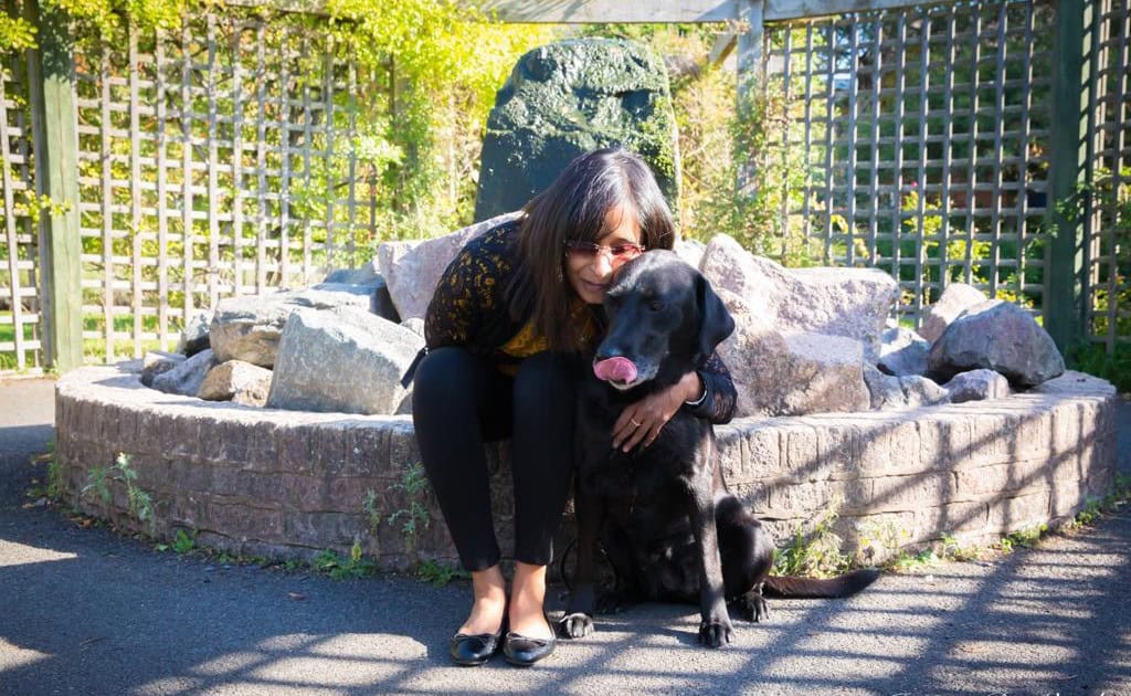 Jaina is sat down on a brick wall, leaning forward to cuddle her black dog Laura. They are posing cheek to cheek, with a green garden behind them.
