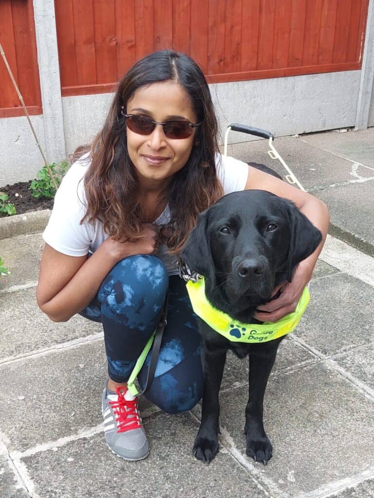 Jaina is kneeling on stone slabs, posing next to her black dog, Kath. The dog has a collar with the words 'Guide Dogs' on it.