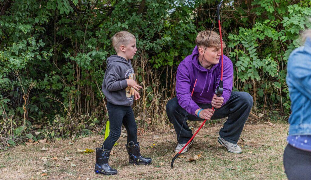 Oakley is stood to the left of James. He has a concentrated face after shooting an arrow at the target. They are both looking to the right and reacting to Oakley's shot.
