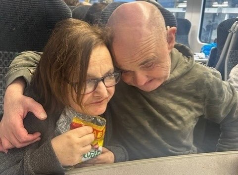 Joe and Joy are sat side by side on a train. Joe has his right arm around Joy's shoulders, and their foreheads are meeting in the middle, touching intimately.