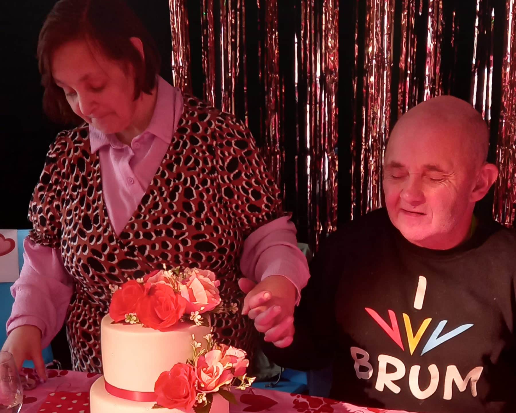 Joe and Joy are standing behind a table with a pink cake on it. They are both holding hands and smiling. Joe has a black jumper on, with icons and words that read 'I love BRUM'