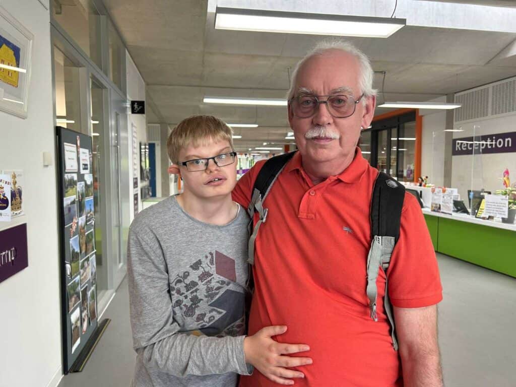A young adult man in a grey jumper stands next to an older man in a red top