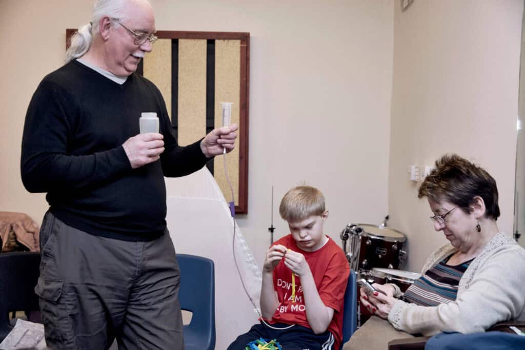 Three people in a room. A man is standing and holding feeding equipment for a young man who is sitting down. A woman sits on the side with her phone in hand.