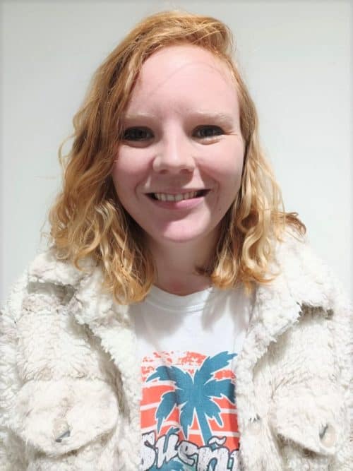 A woman with blonde curly hair wearing a white t-shirt and jacket smiles to camera