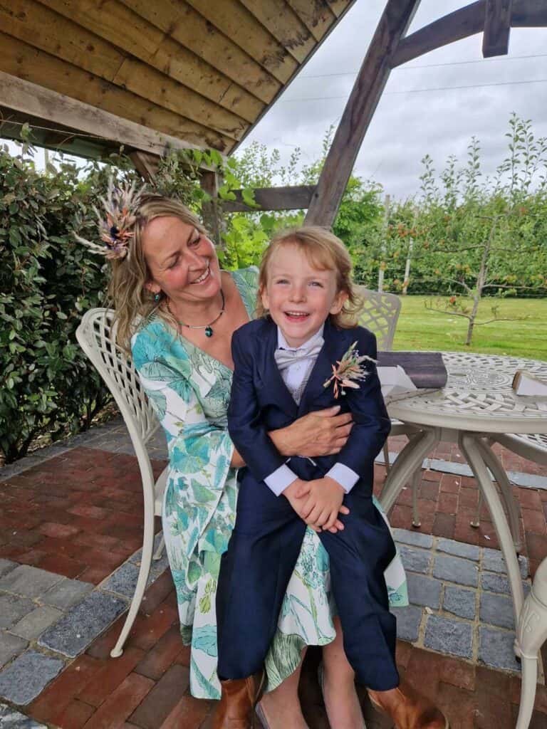 A woman sat with a child on her lap outside. They are dressed smartly and appear to be at a wedding.