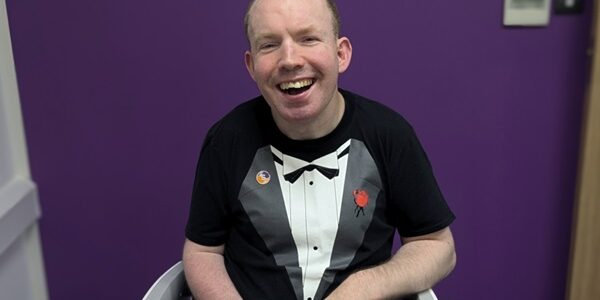 A man wearing a t-shirt with a tuxedo printed on the chest sits in his wheel chair msiling at the camera.
