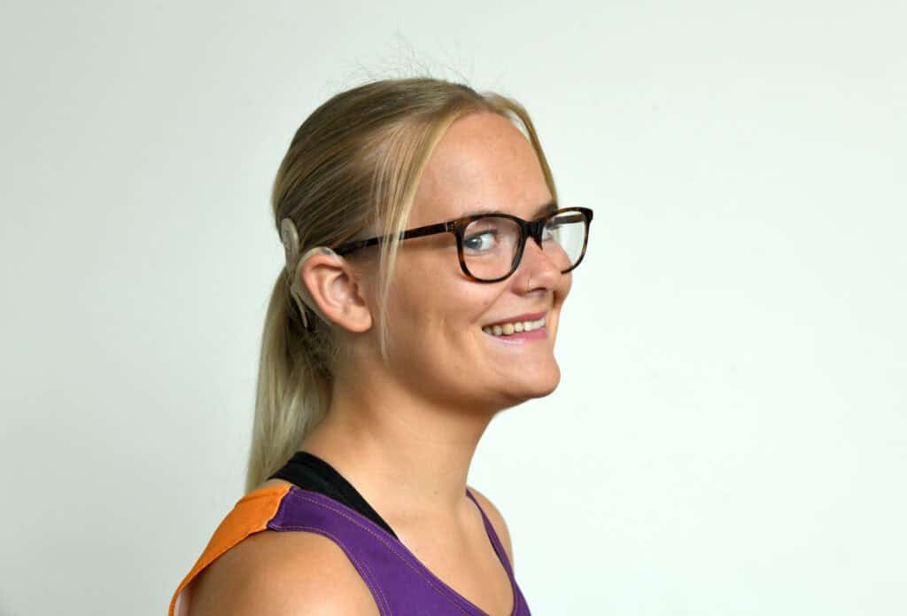 Paige, a white woman with a blonde ponytail and a cochlear implant, smiles at the camera.