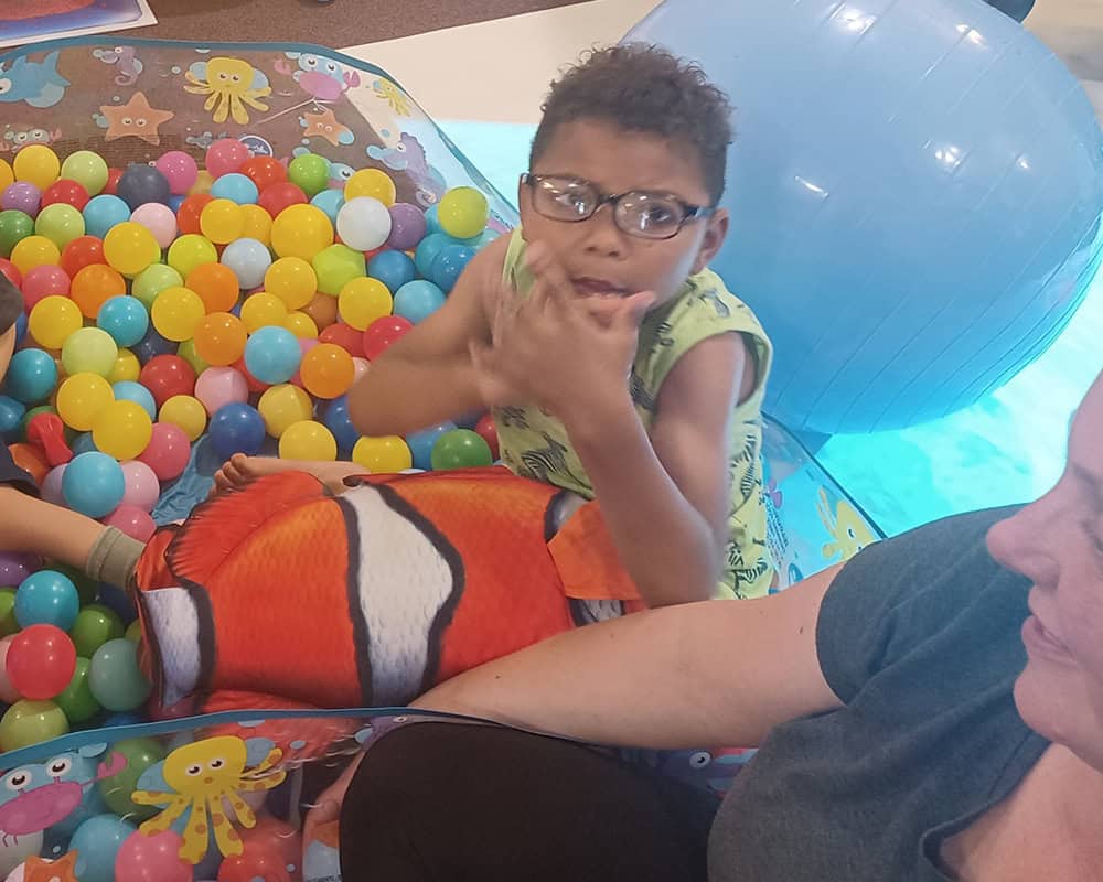 Leo is wearing a green T-shirt and sitting in a ball pit holding a large orange fish. He sits next to his mum Simona and points at the camera.
