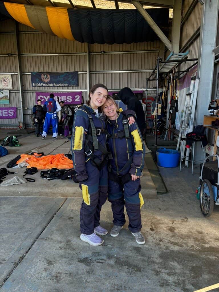 Two women wearing skydiving outfits with their arms around each other, waiting to skydive.