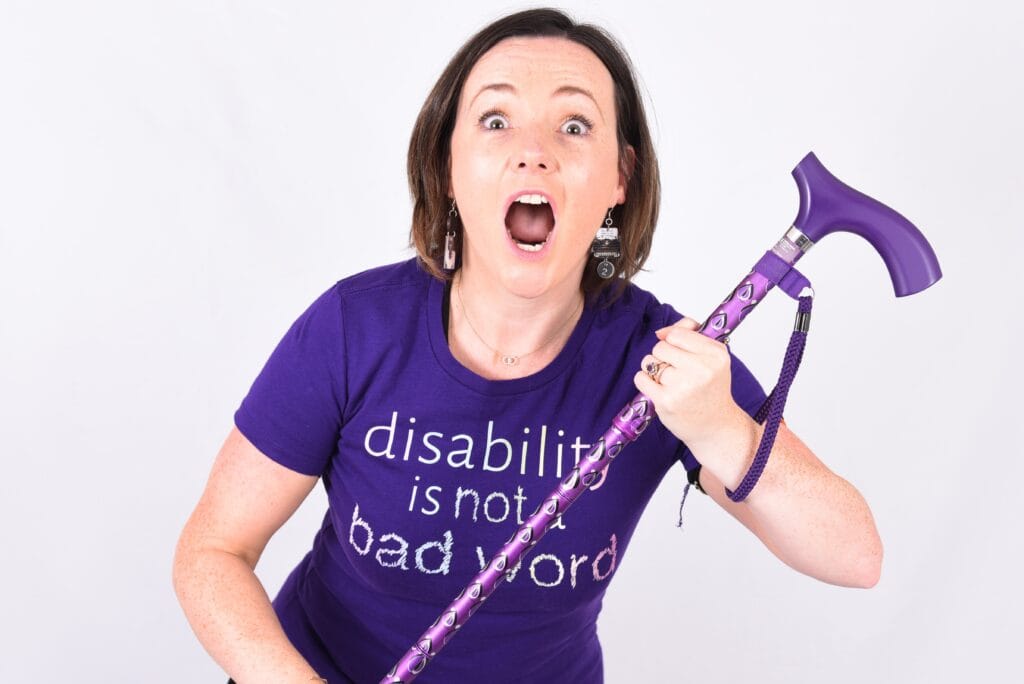 Lisette Auton, a white woman with brown hair, wearing a purple t-shirt saying "disability is not a bad word" and holding a purple mobility aid.