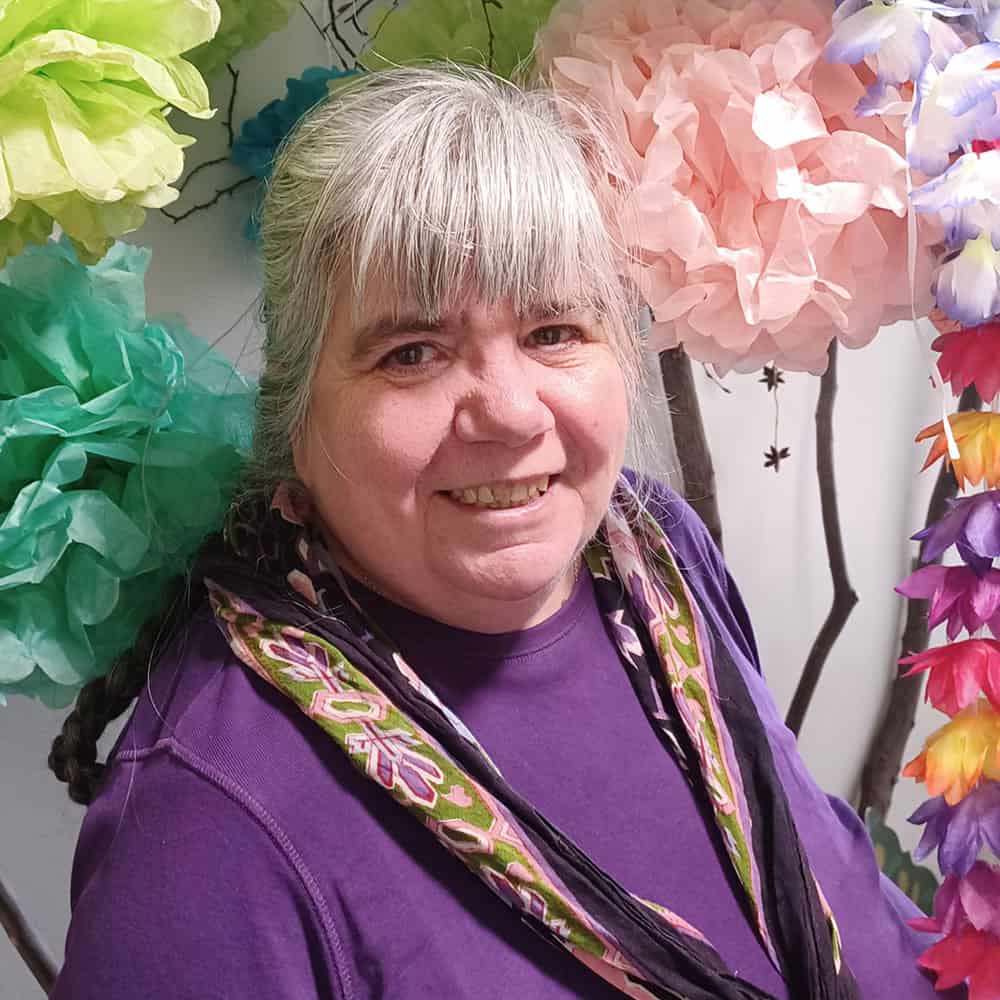 Liz, MSI teacher, is wearing a purple jumper and a scarf. She has grey hair tied back and is smiling at the camera.