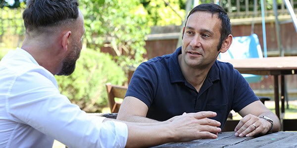 Two men sitting outside at a table talking to each other. / --