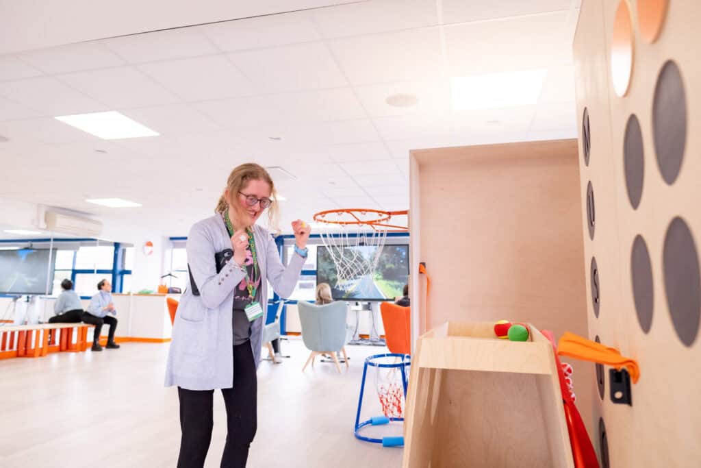 A young lasy with blonde hair tied back raises her fists in celebration. She is standing next to a basket ball hoop in a bright, airy room.