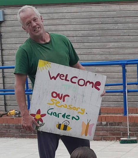 A man holding up a hand painted sign which says Welcome to our sensory garden.