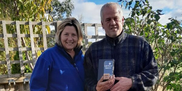 A woman and a man standing in the corner of a garden. The man is holding up a trophy which reads ‘Sense Awards’ on it.