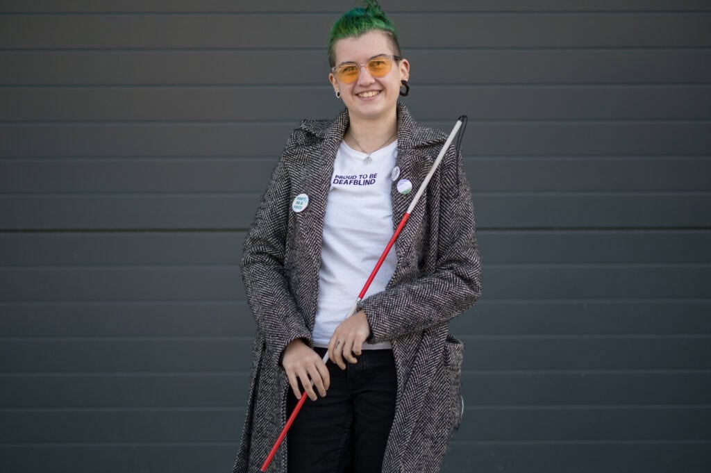 Max, a white genderqueer person with green hair, holding their red and white cane.