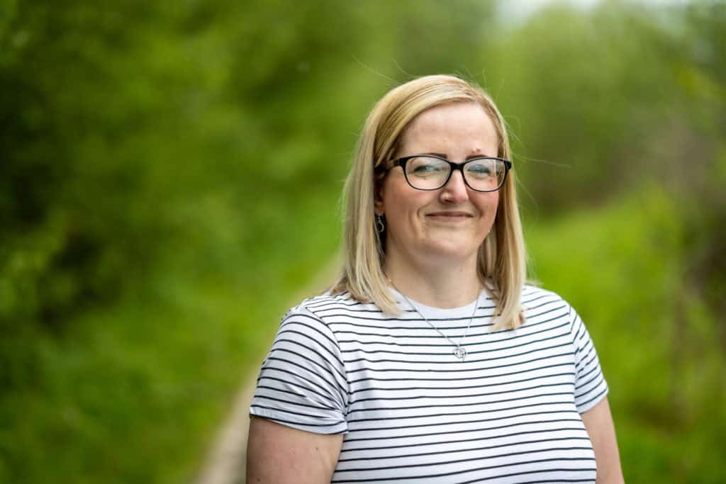 A woman in a stripy top smiles at the camera. There are bushes and grass behind her.