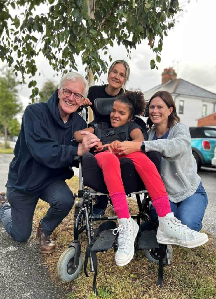 A young person in a wheelchair surrounded by three adults who are holding her hands and the back of the wheelchair.