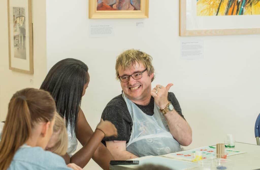 Two people in an art studio smiling and talking. They are wearing aprons.