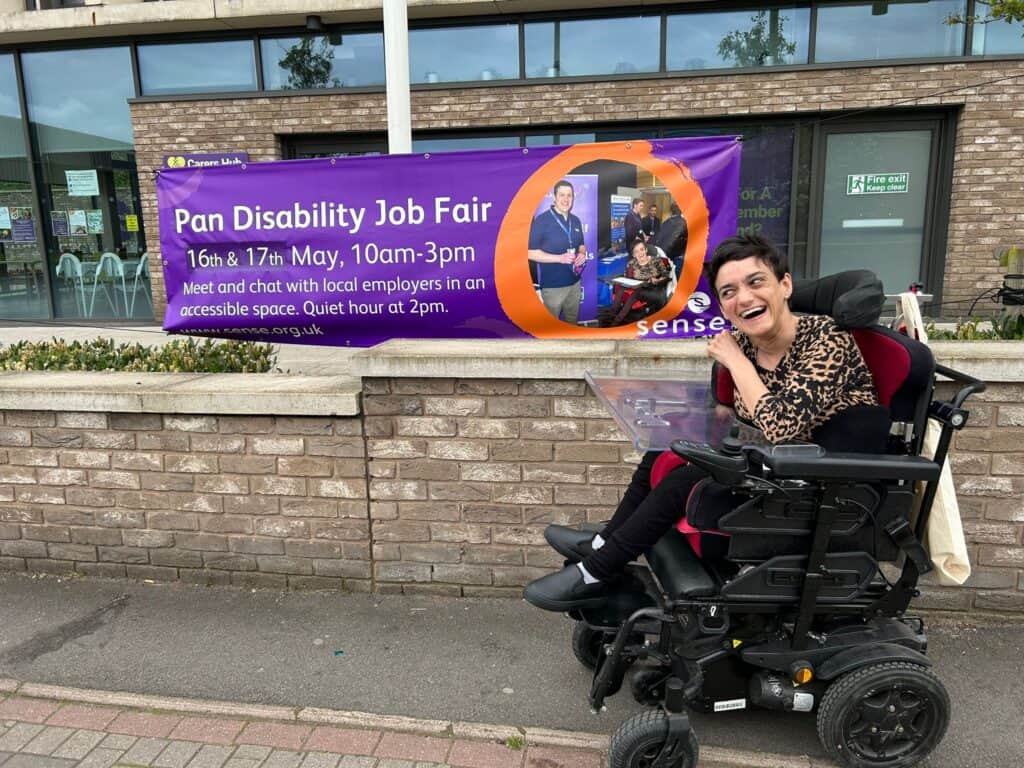 A woman in a wheelchair next to a sign for the Sense Pan Disability Job Fair