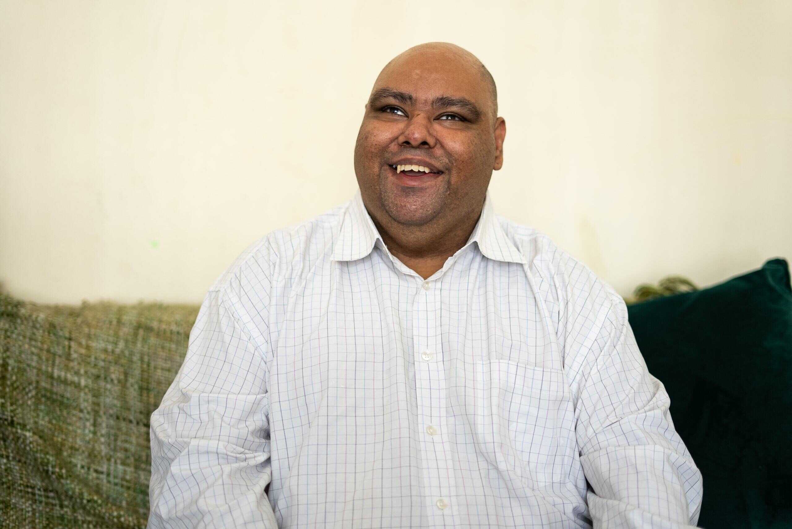 A man in a shirt sits on a sofa inside. He is smiling.