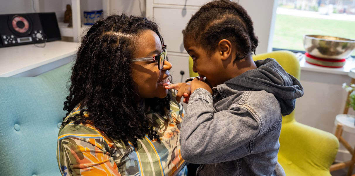 Natalie is facing her son, Azuriah in their living room. They are both smiling widely and staring into each other's eyes. Azuriah has his pinky finger on his mothers bottom lip.