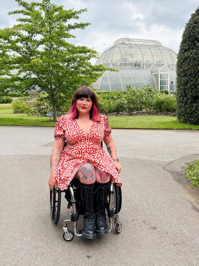 A woman outside with a glass house in the background. She uses a wheelchair and has dyed pink stripes of hair and tattoos on her legs.