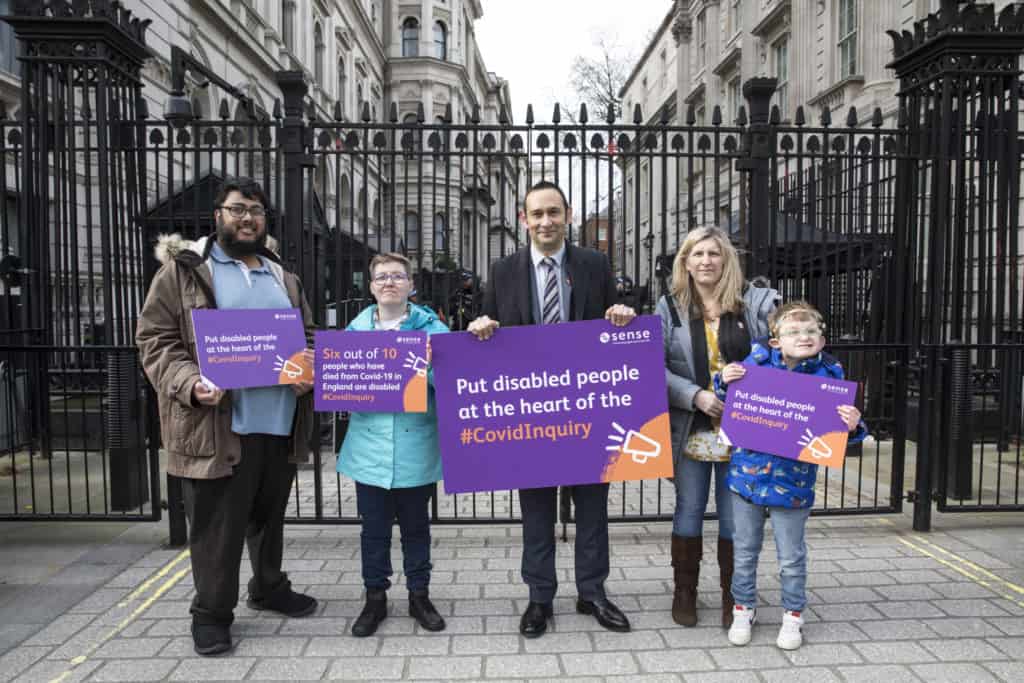 Landscape photo of five people standing in front of black gates outside Number 10, Downing Street. They hold boards showing Sense branding and reading: 'Six out of 10 people who have died from Covid-19 in England are disabled. #CovidInquiry' and 'Put disabled people at the heart of the #CovidInquiry'