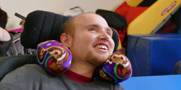 A close-up of a man in a wheelchair and wearing a neck pillow smiling.