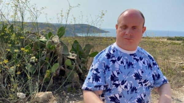 A man wearing a blue and white floral patterned t-shirt sits in front of green plants on a cliff top. Blue sky can be seen meeting the sea in the background.