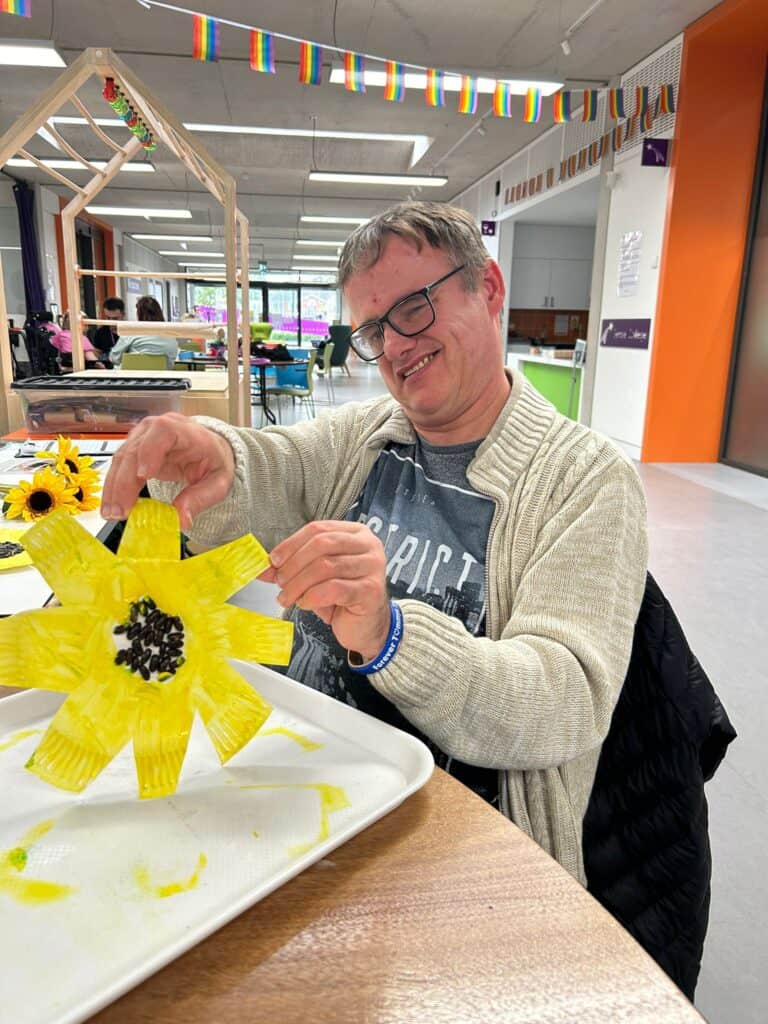 A man sits in Sense TouchBase Pears. He is smiling and holding up a flower he has made using arts and crafts.