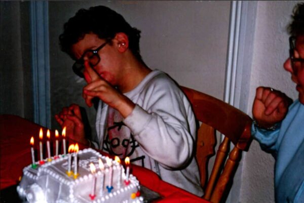 A young man wearing glasses and a white sweatshirt is sitting behind a birthday cake with 12 candles lit.