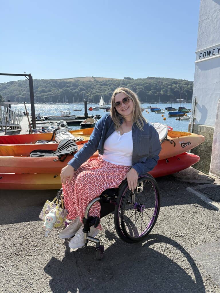 A woman posing in front of water with a pile of canoes behind her. She uses a wheelchair and is wearing sunglasses.