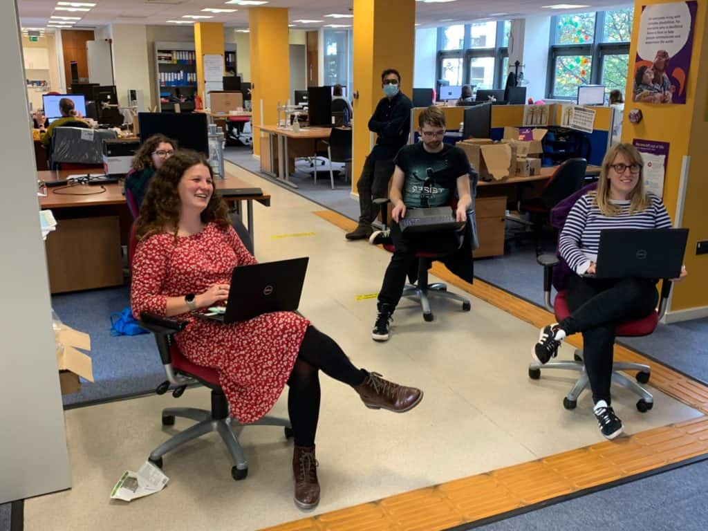 Inside an office, four people sit on office chairs. They have laptops on their laps, and they're looking up at a TV screen. They're smiling excitedl