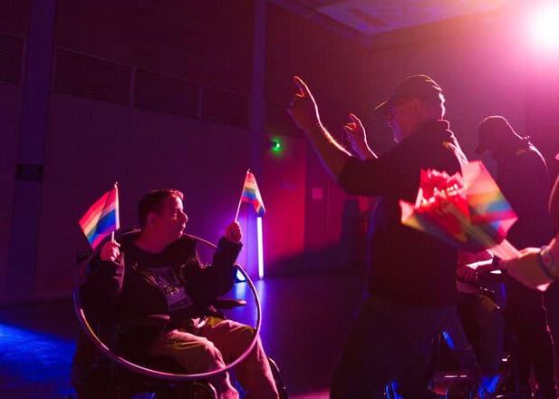 A man in a wheelchair dances, his hands are in the air and he's holding two rainbow flags. Other people are dancing around him