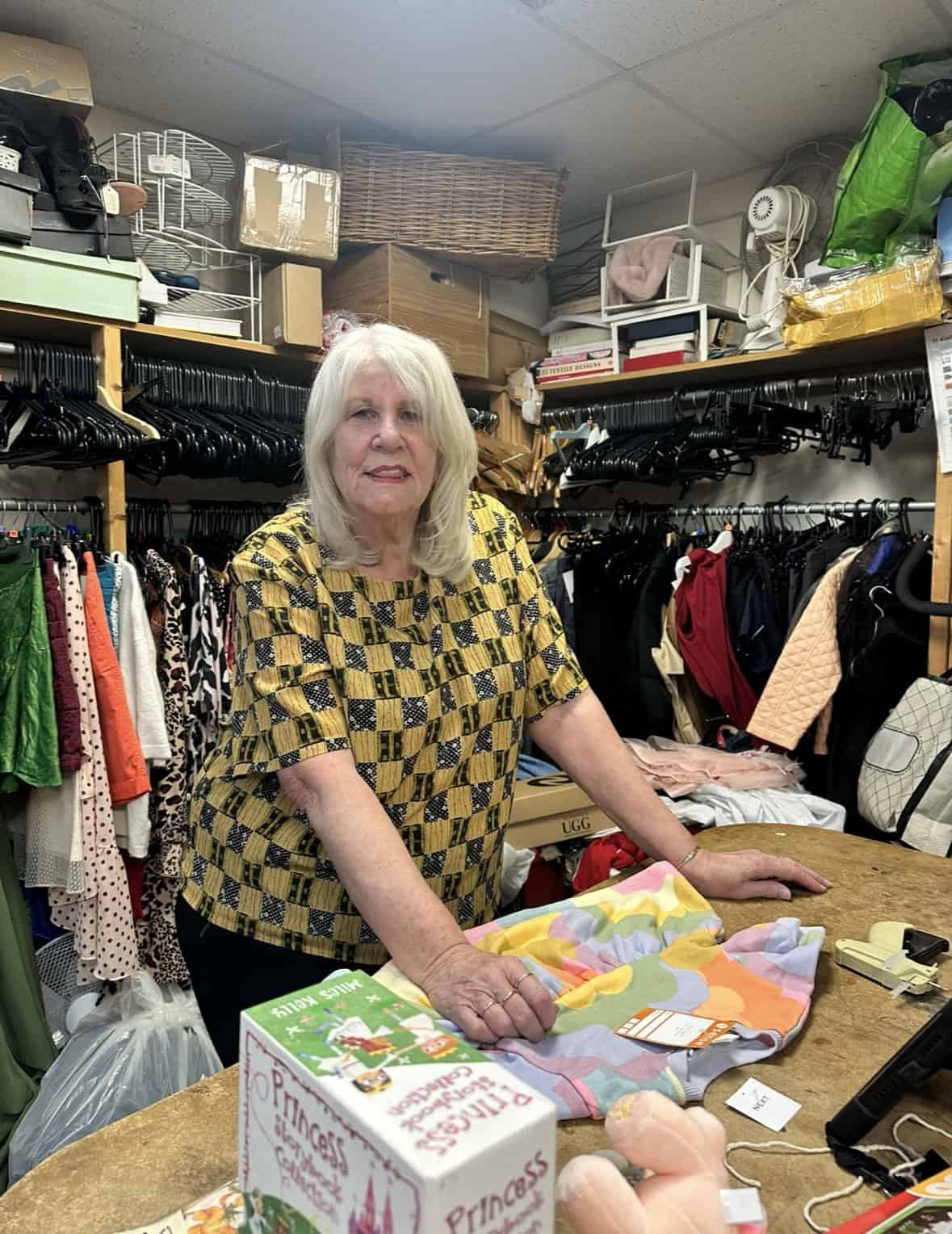 A woman in a charity shop sorting an item. She is surrounded by merchandise.