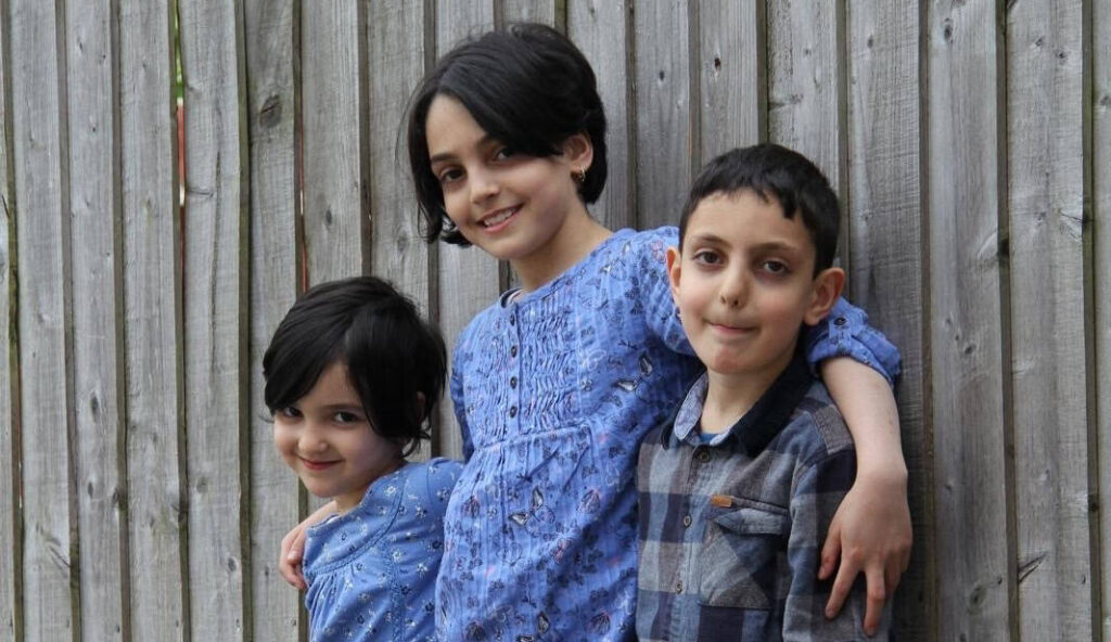 Three siblings stood in a line, all smiling widely. The two girls on the left are in blue dresses and the boy is wearing a chequered shirt.